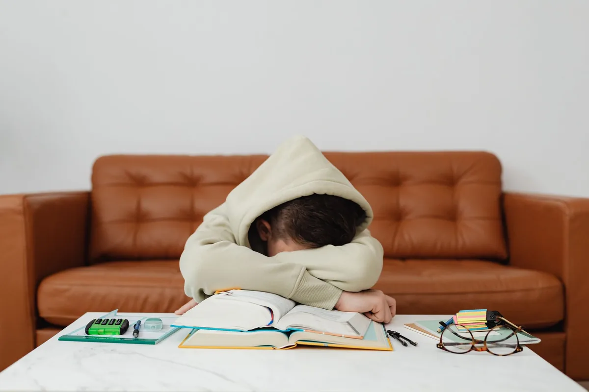 A tired child resting their head on a desk beside an open book and glasses