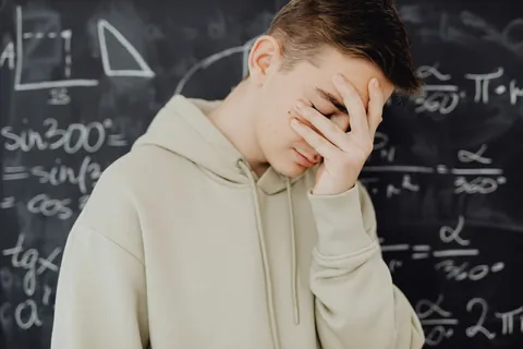 A boy covering his face in frustration in front of a chalkboard full of equations